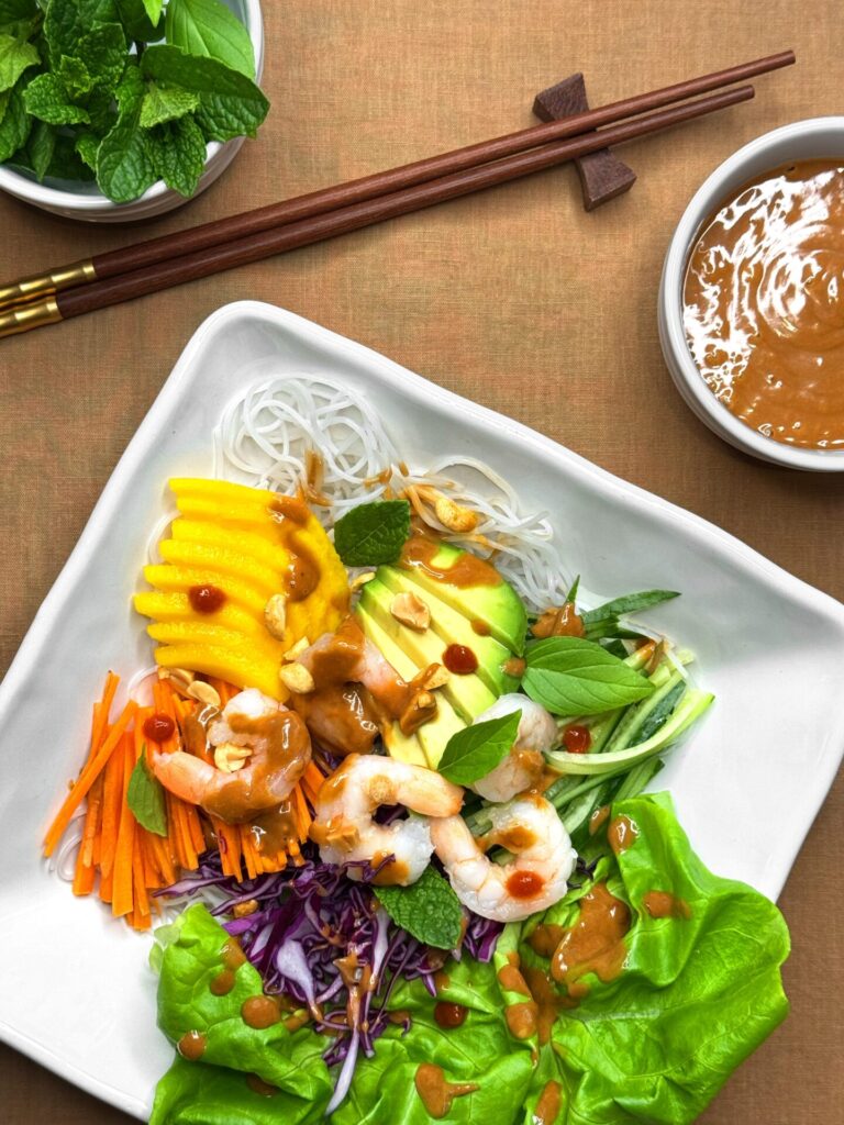 Salade rouleau de printemps déconstruite, avec laitue, crevettes, avocats, carottes, chou, mangue, vermicelles de riz et herbes asiatiques, nappée d'une sauce divine aux arachides.